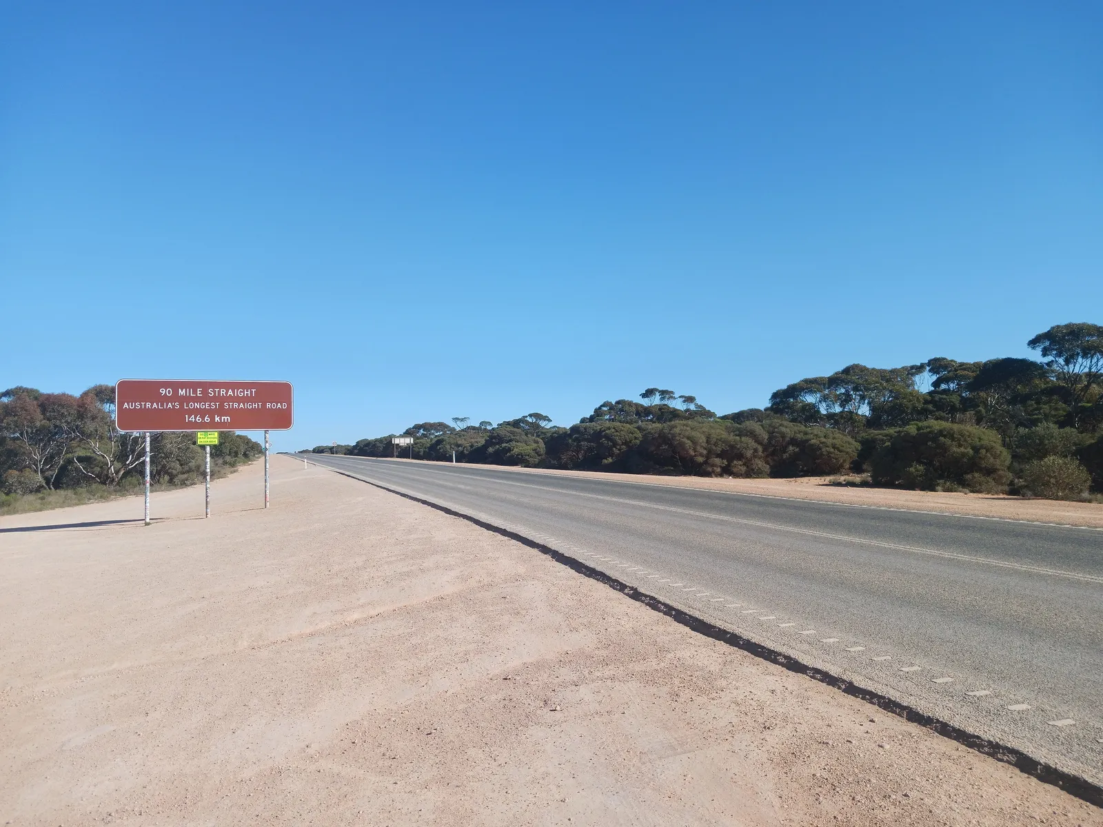 A brown road sign reading 90 Mile Straight, Australia's longest straight road, 146.6 km, beside an empty highway