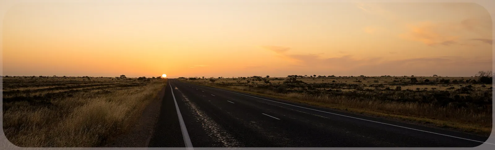 An empty sealed highway running dead straight into a low orange sun across the Nullarbor Plain