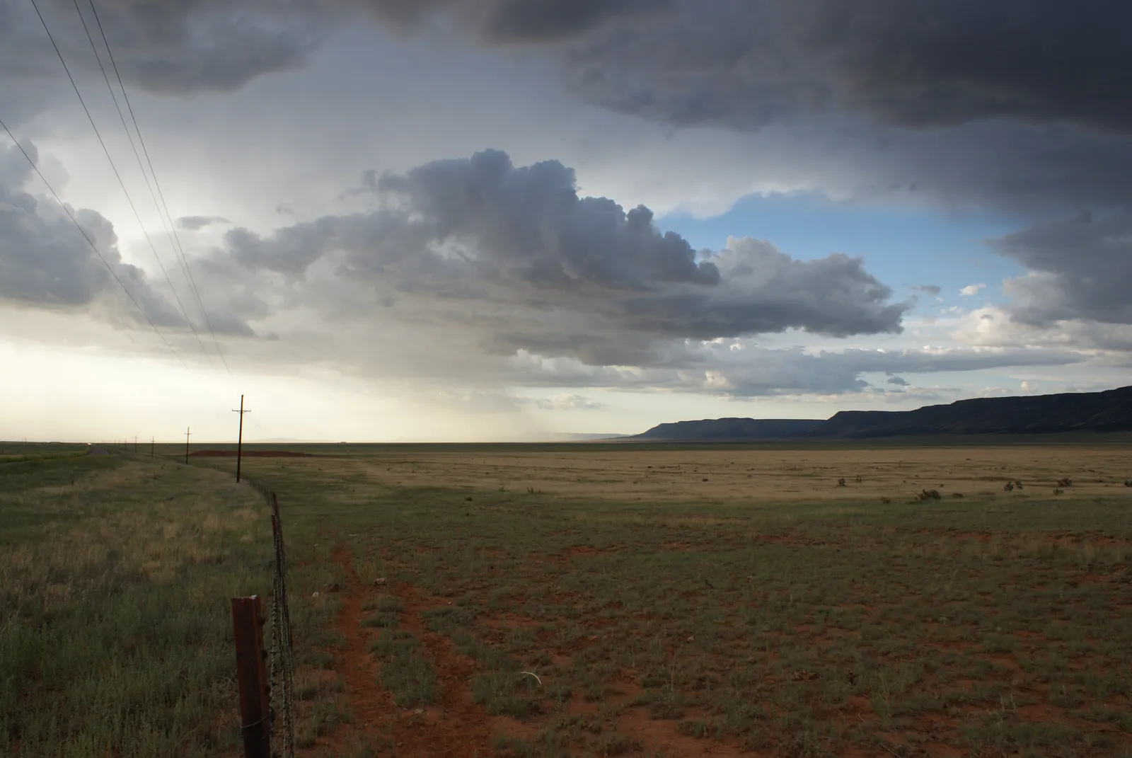 A storm cloud over open grassland, with power lines beside a fence and a flat-topped mesa in the distance