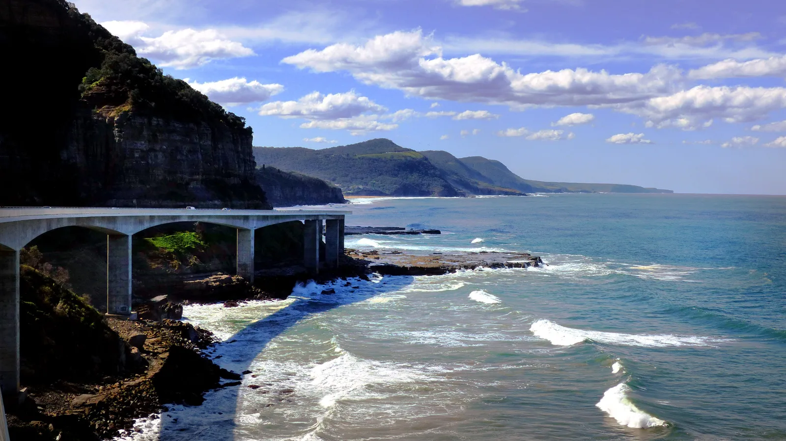 A concrete bridge curving along a sea cliff above the Tasman Sea, under a blue sky with scattered cloud